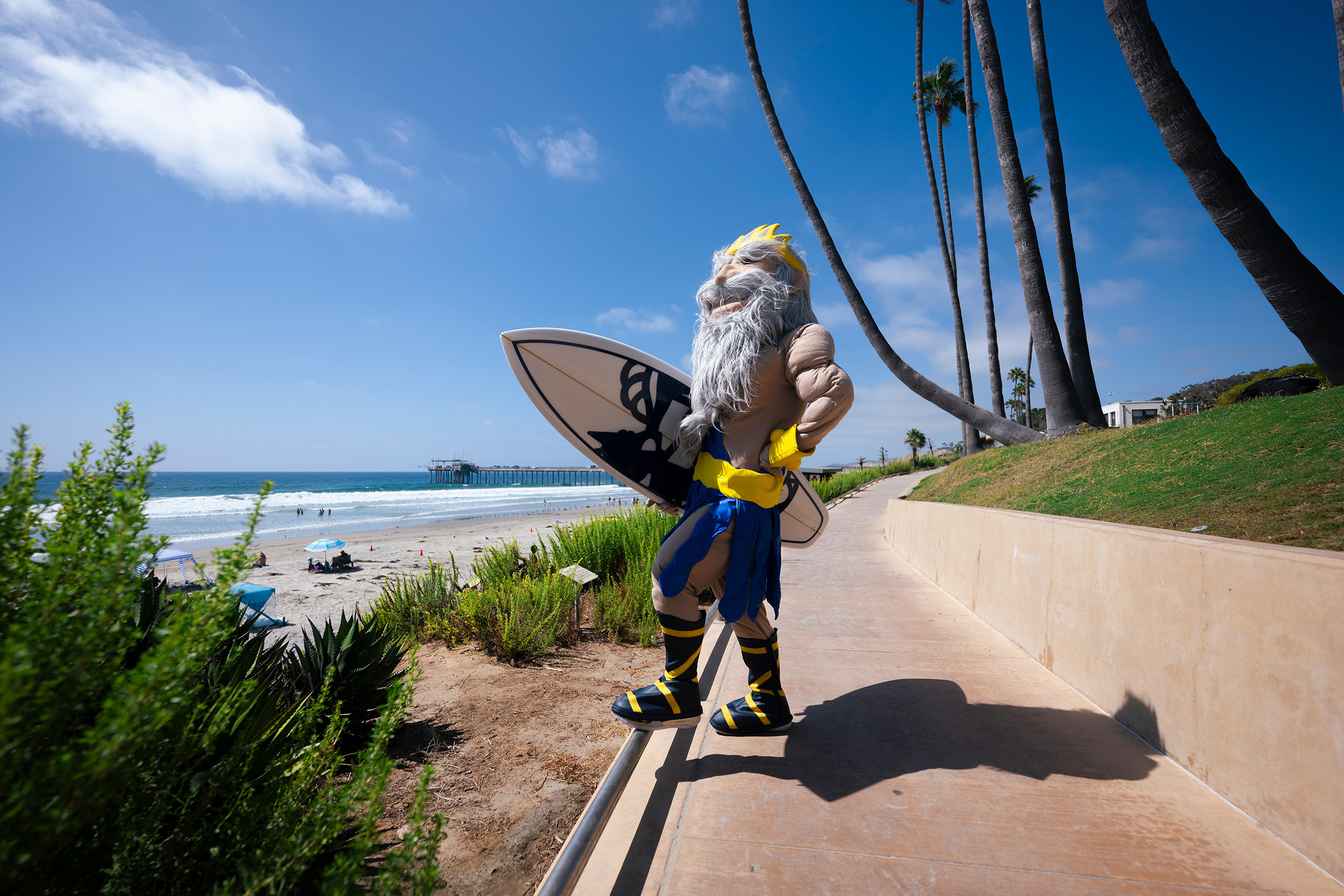 King Triton mascot holds surfboard while gazing at ocean.