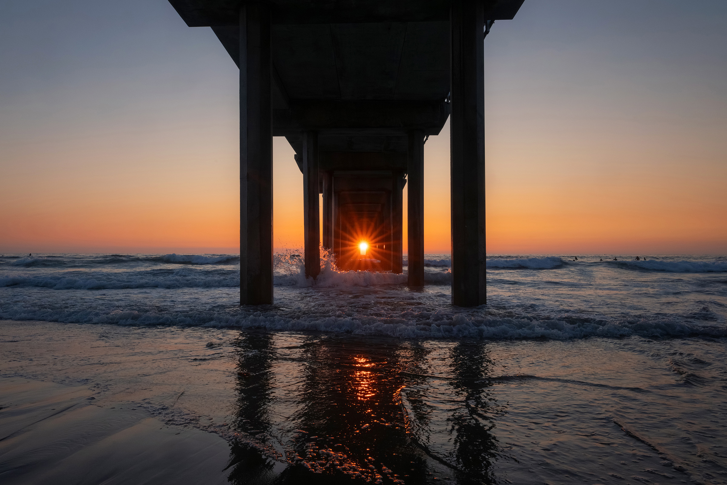 The sunset shines between the pillars beneath a pier, just above the ocean horizon.